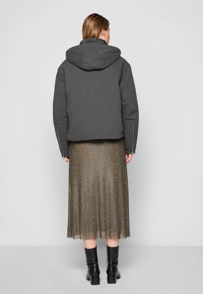 Dunkelgraue, gefütterte Jacke mit Kapuze und cropped Design. Kombiniert mit einem plissierten, metallisch goldenen Rock und schwarzen Ankle Boots.