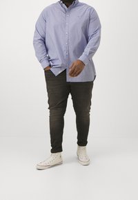 Man staat in een blauw geruit overhemd met knopen, donkere slim fit spijkerbroek, witte hoge sneakers, met één hand in zijn zak.