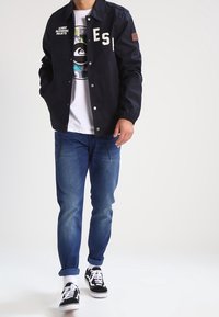 Chaqueta azul marino con letras blancas, corte relajado, combinada con una camiseta gráfica blanca y vaqueros azules. Zapatillas negras y blancas completan el conjunto.