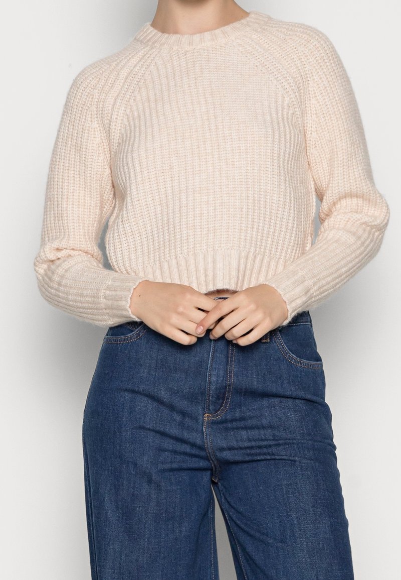 Femme portant un pull en tricot crème et un jean taille haute bleu foncé, mains jointes à la taille.