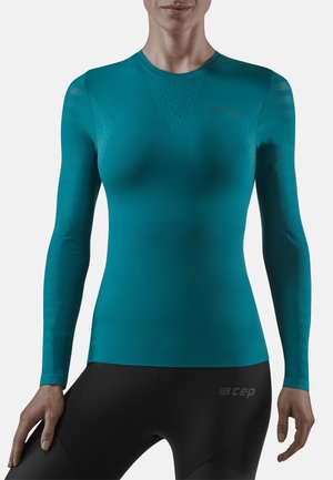 Personne portant un haut de sport ajusté à manches longues couleur sarcelle et un legging noir avec des logos de marque, debout avec les bras légèrement pliés sur les côtés.
