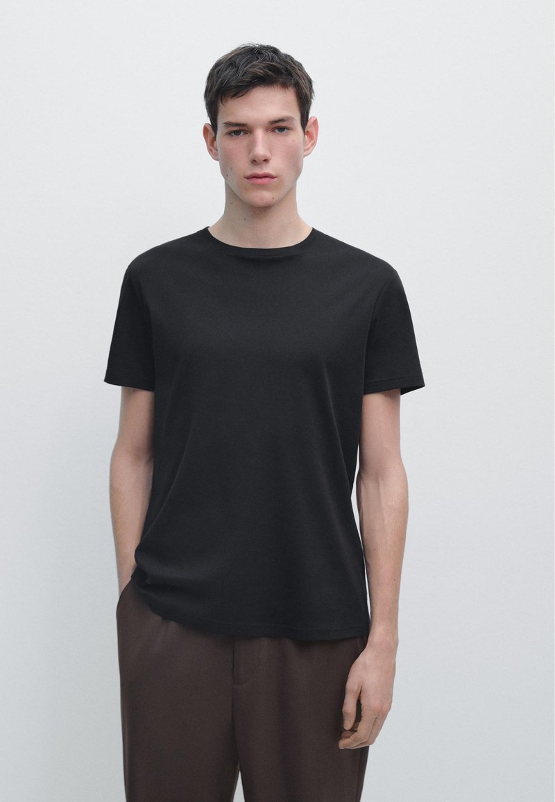 Junger Mann mit kurzen dunklen Haaren, der ein einfaches schwarzes T-Shirt und braune Hosen trägt, vor einem weißen Hintergrund stehend.