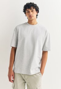 Jasnoszara bawełniana koszulka z okrągłym dekoltem i krótkimi rękawami, o luźnym kroju i subtelnej fakturze. Noszona z beżowymi szortami typu cargo.