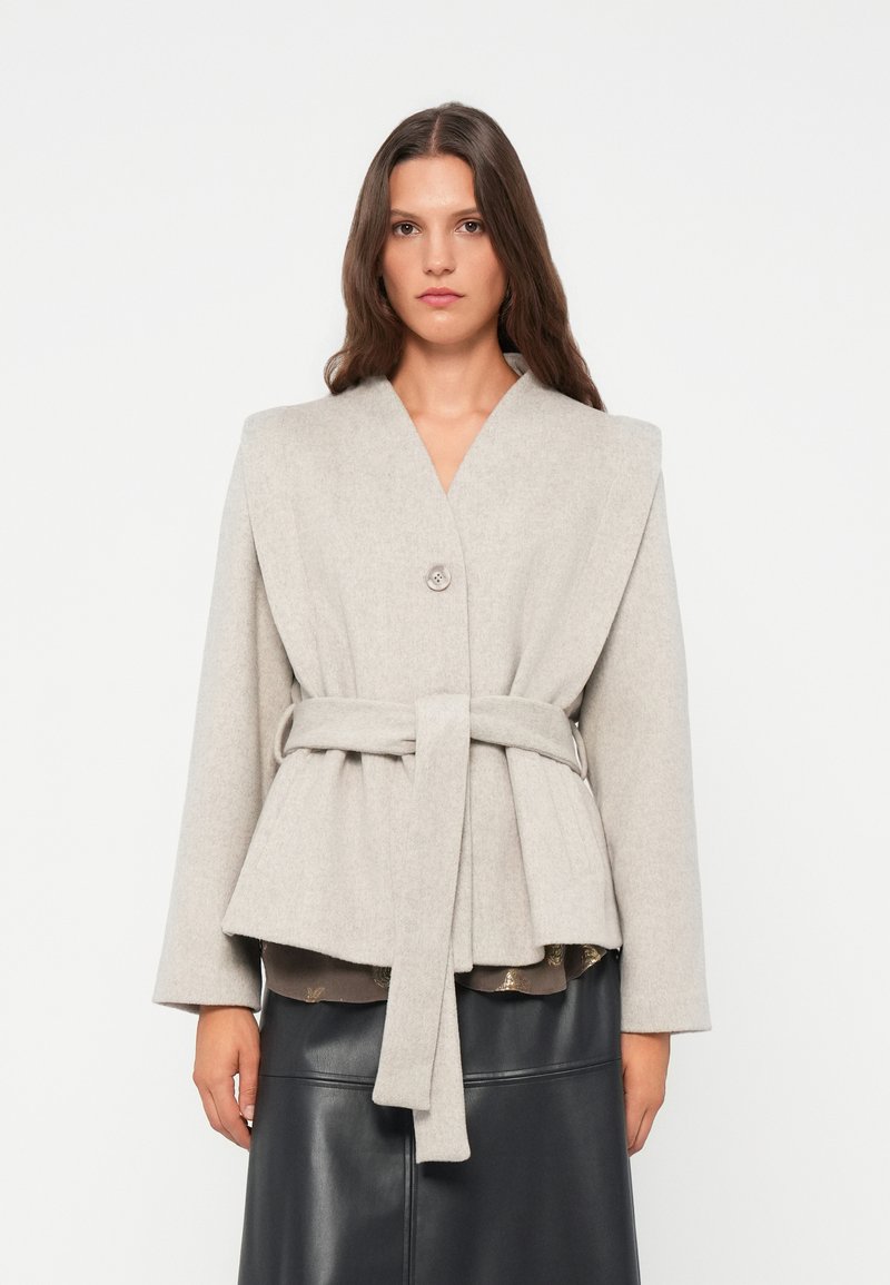 Giacca di lana grigia chiara con ampio risvolto, chiusura con un solo bottone e vita cintrata, caratterizzata da una silhouette peplum e maniche lunghe.