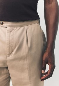 Beige maßgeschneiderte Hose mit vorderen Bundfalten, einem taillierten Bund mit Knopf und Seitentaschen. Glatter Stoff, vertikale Struktur und eine figurbetonte Passform.