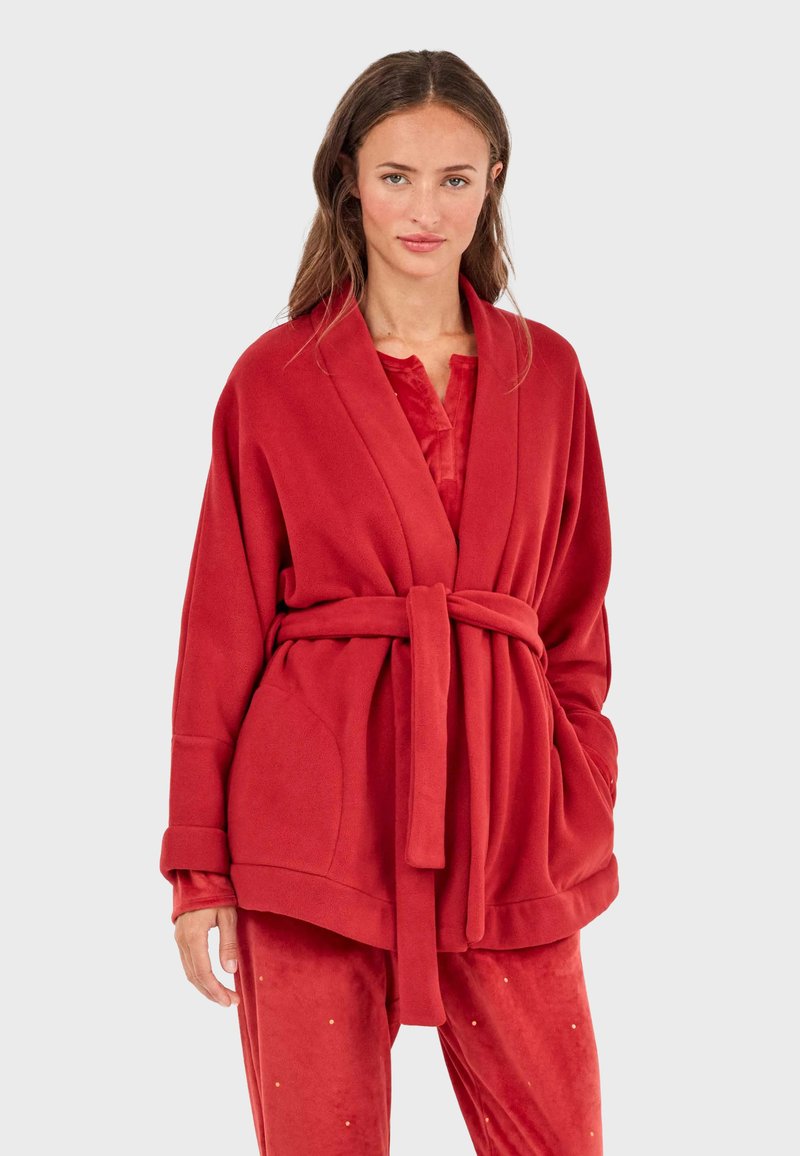 Chaqueta de forro polar rojo con cinturón, mangas extragrandes y bolsillos laterales. Presenta una textura suave y un acabado liso, combinada con pantalones a juego.