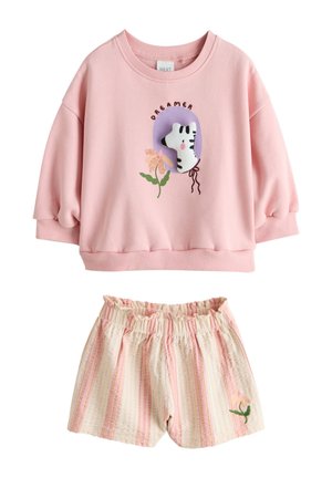 Kind mit lockigem Haar in einem rosa Pullover und weißem Rock läuft auf Gras in einem sonnigen Außenpark mit Bäumen im Hintergrund.