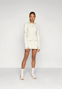 Heled kreemikas kudumiga zippažakk, millel on pikad varrukad ja võrgumaterjalist aktsendid. Koos sobiva seeliku ja beežide sportlike kingadega.