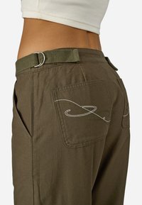 Tamsiai žali medvilniniai kelnių su šoninėmis ir galinėmis kišenėmis, papuošti baltais ir žaliais siuvinėjimais bei sidabriniu D formos diržo kilpu.