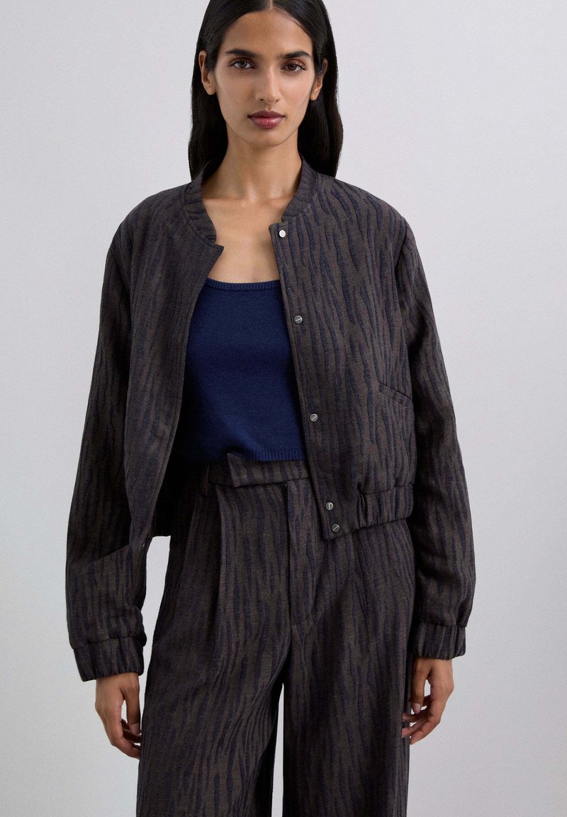 Chaqueta bomber de patrón oscuro con cuello redondo, cierre de botones y puños de canalé, combinada con una blusa de tirantes ajustada en color navy.