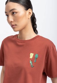 Maglietta rossa in cotone con tre piccole applicazioni grafiche: un quadrato giallo, una chiave verde e una figura verde, disposte nella zona del petto.