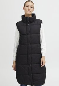Schwarze Steppweste mit hohem Kragen, versehen mit Druckknöpfen, einem gesteppte Design und mittellanger Schnitt, getragen über einem weißen Pullover.