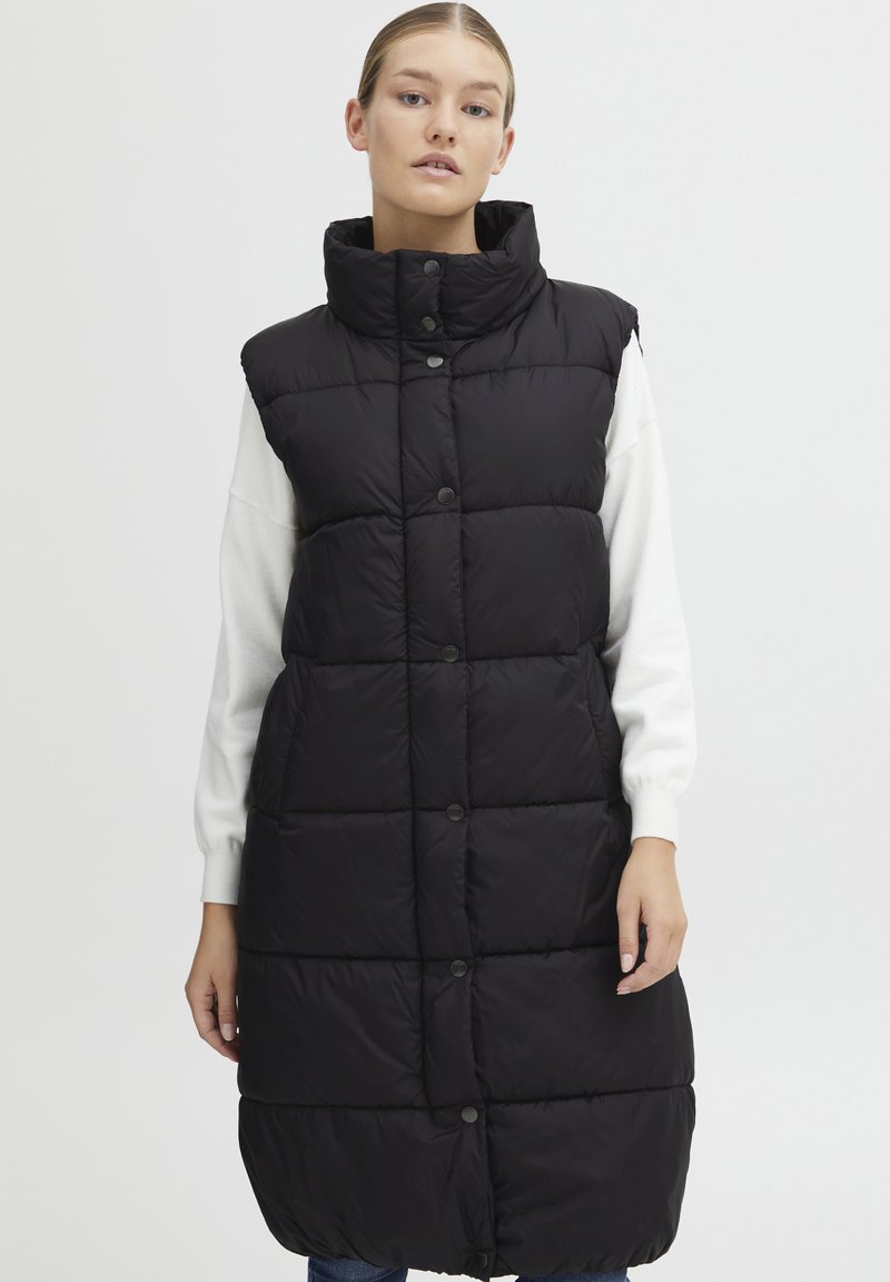 Schwarze Steppweste mit hohem Kragen, versehen mit Druckknöpfen, einem gesteppte Design und mittellanger Schnitt, getragen über einem weißen Pullover.