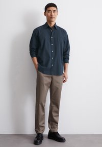 Dunkelblaues Hemd mit strukturiertem Muster, kombiniert mit beigefarbenen Hosen und schwarzen Schuhen, vor einem schlichten Hintergrund stehend.