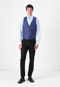 Colete azul texturizado com cinco botões, usado sobre uma camisa azul clara, combinado com calças pretas e sapatos pretos, em pé contra um fundo neutro.