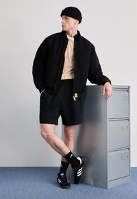 Black bomber jacket with a beige shirt, black shorts, and black sneakers with white stripes. Room with gray filing cabinet and blue floor.
