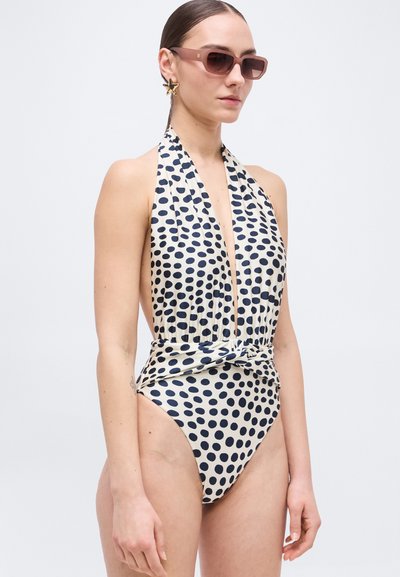 Žena v bezrukávových plavkách s bodkami a uzlom na krku, hviezdicové náušnice a obdĺžnikové slnečné okuliare.