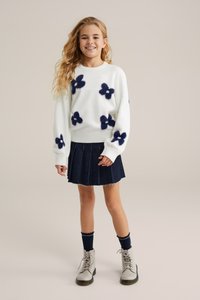 Pulover alb cu modele florale pufoase în nuanțe de navy, asortat cu o fustă plisată din denim închis. Modelul poartă cizme gri cu șireturi și sosete navy.