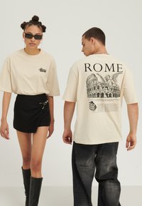 Two models wear beige t-shirts with "ROME" and Colosseum prints; woman has black skirt and boots, man wears dark jeans and chain necklace.