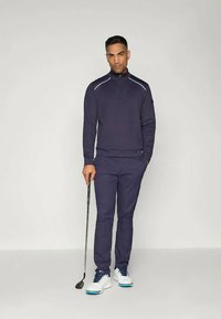 Jūras spēku golfam paredzēta puloveris ar augstu apkakli un baltām akcentēm, saskaņots ar atbilstošām biksēm un baltām golfa kurpēm. Vienā rokā turēts golfa nūja.