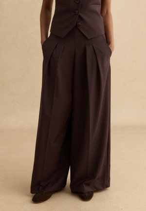 Persona che indossa pantaloni plissettati a gamba larga color marrone scuro con tasche laterali e gilet abbinato con bottoni, in piedi con le mani nelle tasche.