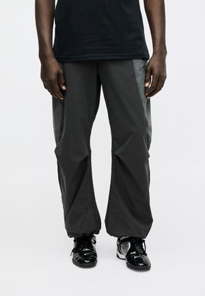 Person iført løstsiddende mørkegrå bukser, sort T-shirt og skinnende sorte snøresneakers stående mod en hvid baggrund.