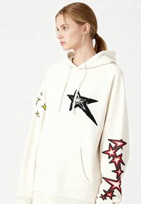 Felpa con cappuccio color crema con tasca frontale, caratterizzata da grafiche di stelle nere e rosse sul corpo e sul braccio, e dettagli di stelle gialle sulla manica.