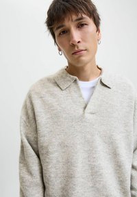 Lys grå uldsweater med faldende hals, afslappet pasform og synlig tekstur. Modellen har en hvid t-shirt på under.