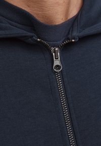 Mørkeblå zip-up hoodie med en glat tekstur, der har en halv-lynlås lukning og en ribbet krave. Metal lynlåselementer er synlige.