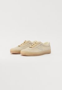 Beige semskosneakers med en rund form, kontrasterende lys tan snørebånd og en teksturert gummisåle. Har minimal søm og logo detaljer.
