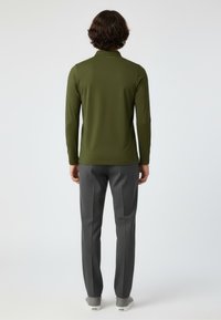 Mann, der mit dem Rücken zur Kamera steht und ein olivgrünes langärmliges Hemd, graue Hose und graue Slipper trägt, vor einem schlichten Hintergrund.