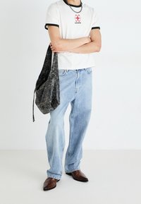 Persona vestida con una camiseta blanca con ribete negro, jeans de color azul claro, zapatos marrones de punta, sosteniendo un bolso de hombro negro texturizado.