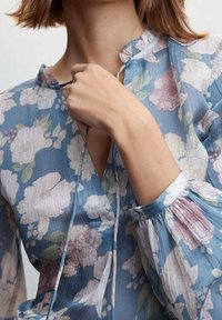 Blomstret bluse i lyseblå med flæsekrave, lange ærmer og samlede manchetter, med hvid, pink og grøn blomstret mønster.