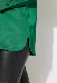 Zielona satynowa koszula z zaokrąglonym dołem i bocznym rozcięciem, w połączeniu z czarnymi skórzanymi spodniami. Materiał wydaje się gładki z delikatnym połyskiem.