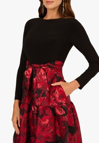 Schwarzes langärmliges Oberteil mit einem Rock in rot-schwarzem Blumenmuster, ausgestattet mit einem gebundenen Taillenbund und Taschen, glatter Textur und geschichteter Gestaltung.