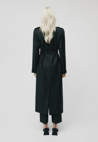 Zwarte op maat gemaakte jas met een ceintuur in de taille, lange mouwen en een achtercut-out, gecombineerd met kortere zwarte broeken en schoenen met blokhak.