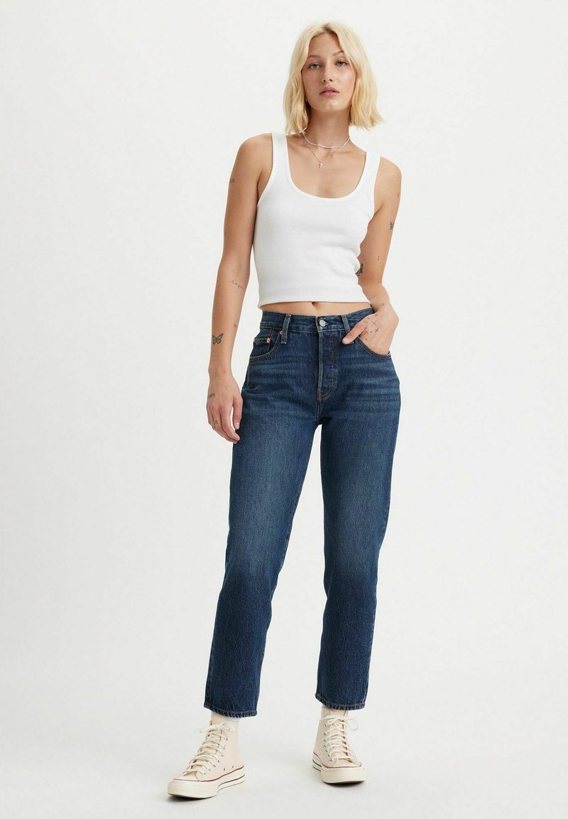 Jeans scuri a vita alta con cinque tasche, con una vestibilità dritta e dettagli di cucitura sottili, abbinati a un top a maniche corte bianco a coste e sneakers.