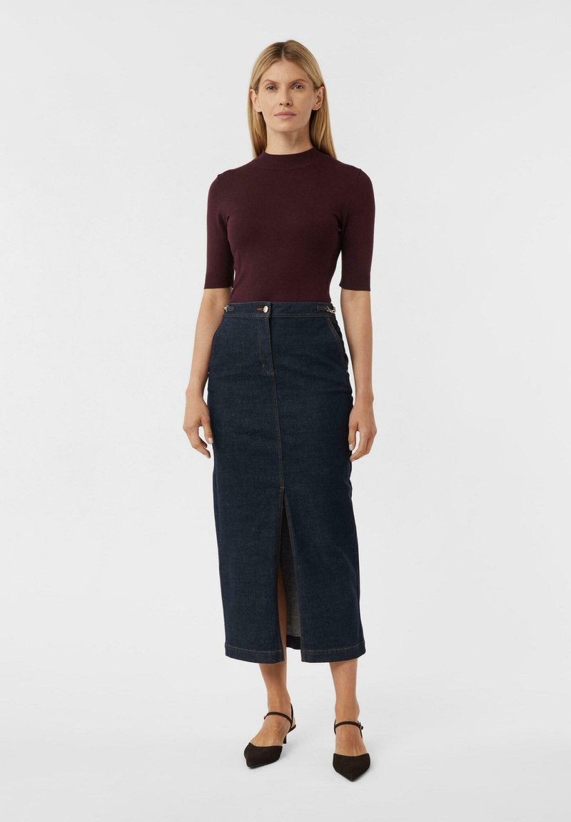 Frau in einem dunkelpurpurfarbenen kurzärmeligen Oberteil, hoch taillierter schwarzer Jeans-Midirock mit Schlitz vorne und schwarzen spitzen Riemchen-Schuhen, die steht.