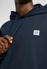 Sudadera azul marino hecha de tejido texturizado. Presenta un bolsillo frontal, cordones y una etiqueta blanca con bordes azules en el pecho.