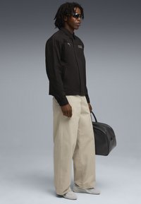 Sort zip-up jakke med logo, beige brede bukser, grå sneakers og sort duffel bag. Funktioner afspejler logoer og forskellige teksturer.