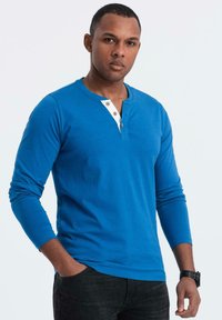 Maglietta a maniche lunghe henley blu con un colletto bianco e bottoni in legno. Presenta un design aderente e una texture morbida. Abbinata a jeans scuri.