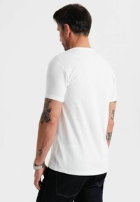Camiseta blanca de manga corta hecha de algodón, con cuello redondo y un corte holgado. El modelo tiene tatuajes visibles en los brazos.