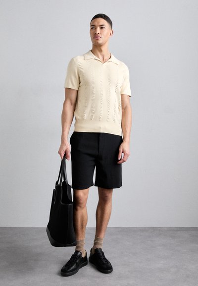 Kortærmet cremefarvet strikpolo med strukturerede lodrette striber, parret med sorte shorts og sorte sneakers. Holder en sort tote bag.