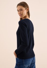 Marineblaues Langarmshirt mit rundem Halsausschnitt, glattem Stoff, taillierter Silhouette und einem dezenten Saumdetail, kombiniert mit Denim-Jeans.