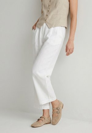 Kobieta w beżowej kamizelce zapinanej na guziki, białych szerokich spodniach i beżowych zamszowych płaskich butach z podwójną klamrą, stojąca na jednolitym tle.