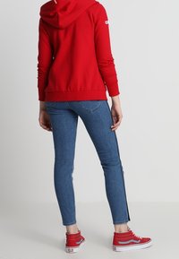 Rode hoodie gemaakt van zacht stof, aangesloten model met een capuchon. Draagt daarbij blauwe slim-fit jeans met zijstrepen en rode sneakers.