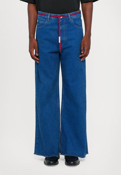 Széles lábú farmernadrág kék denim anyagból, nyers szegéssel, piros húzózsinórral és ellentétes varrásokkal. A modellen fekete lábbeli van.