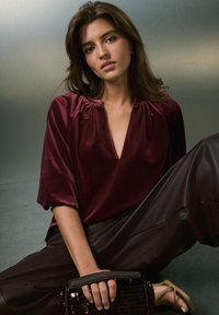 Haut en velours bordeaux avec un décolleté en V profond et des épaules froncées, associé à un pantalon large en cuir marron. Sac à main noir texturé à la main.
