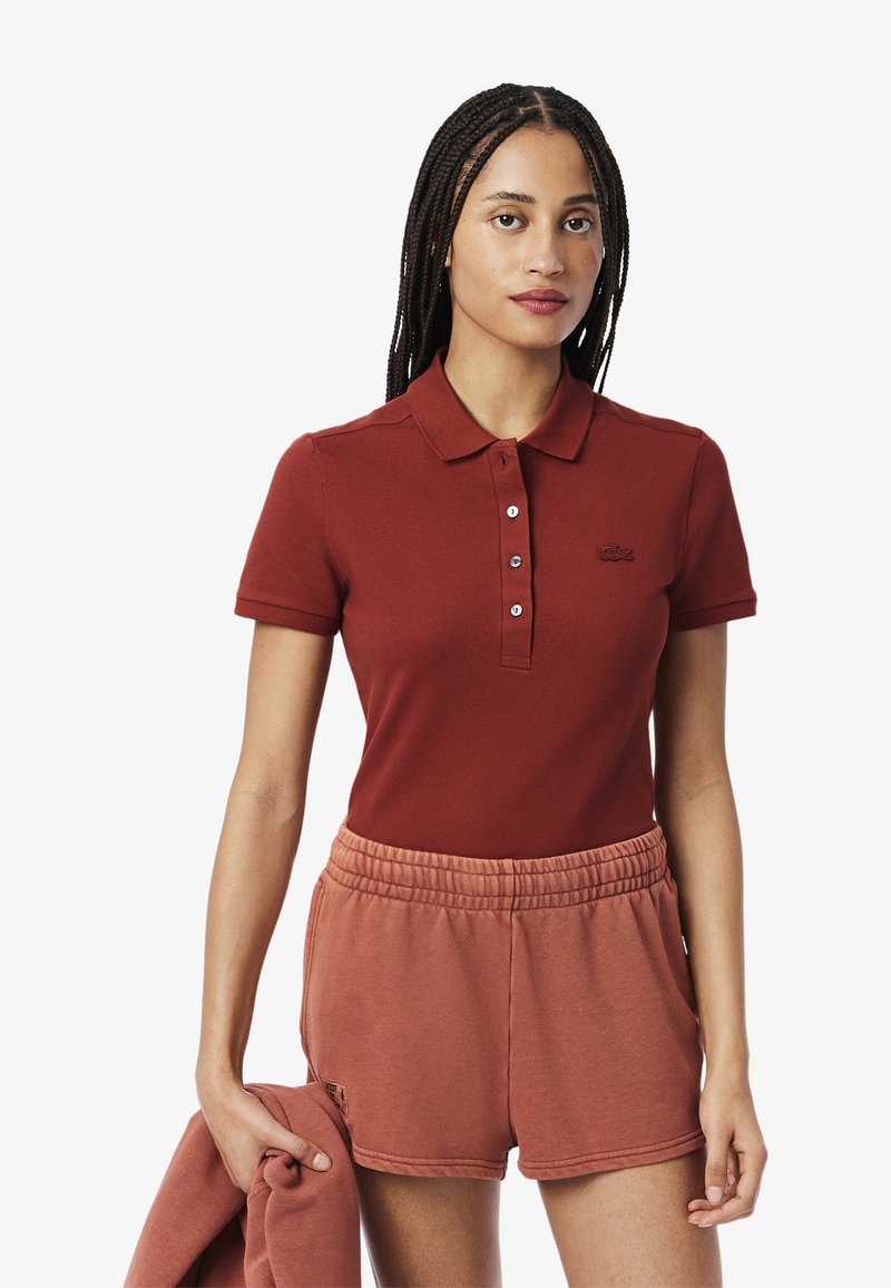 Punainen polo-paita, jossa on kaulus, kolme nappia ja brodeerattu logo. Yhdistetty vaaleanpunaisiin shortseihin, joissa on joustava vyötärö. Sileä pinta.