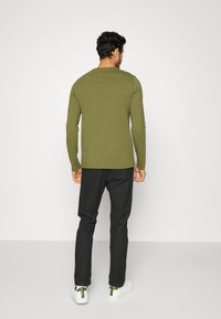 Camisola de manga comprida verde-oliva, design ajustado, combinada com calças pretas e sapatilhas brancas com detalhes verdes, fundo simples.